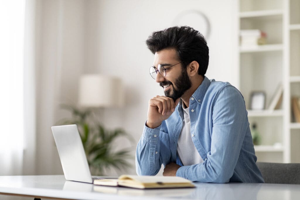 Handsome Indian Male Writer Sitting At Desk With Laptop And Smiling